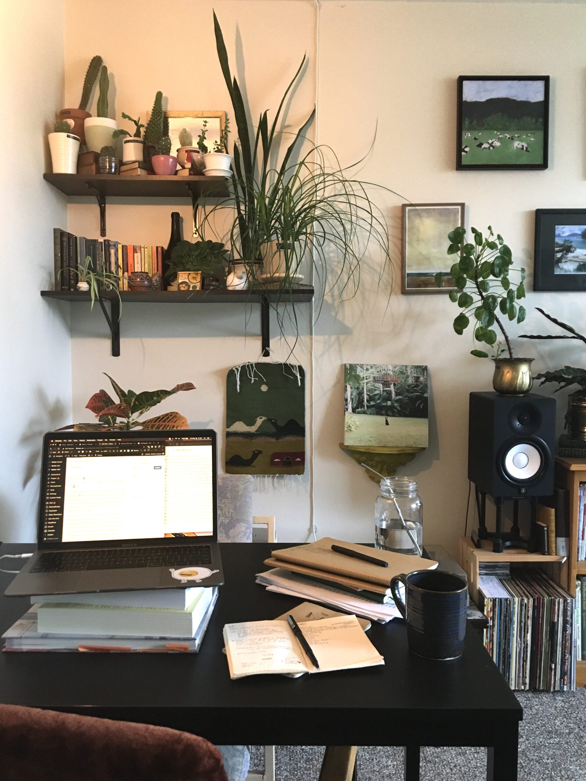 A laptop, notebooks, and mug of coffee sit on a desk in front of a wall of plants and books