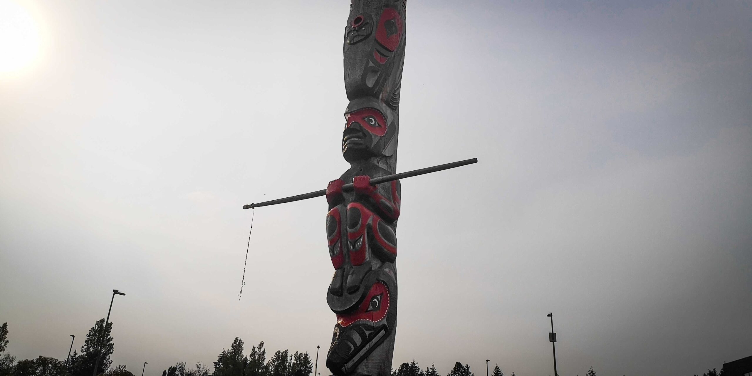 The photo frames Heeshka a tlaquiaht, a whaler who is holding a wooden harpoon. The pole is brown with red and black designs which shape the images