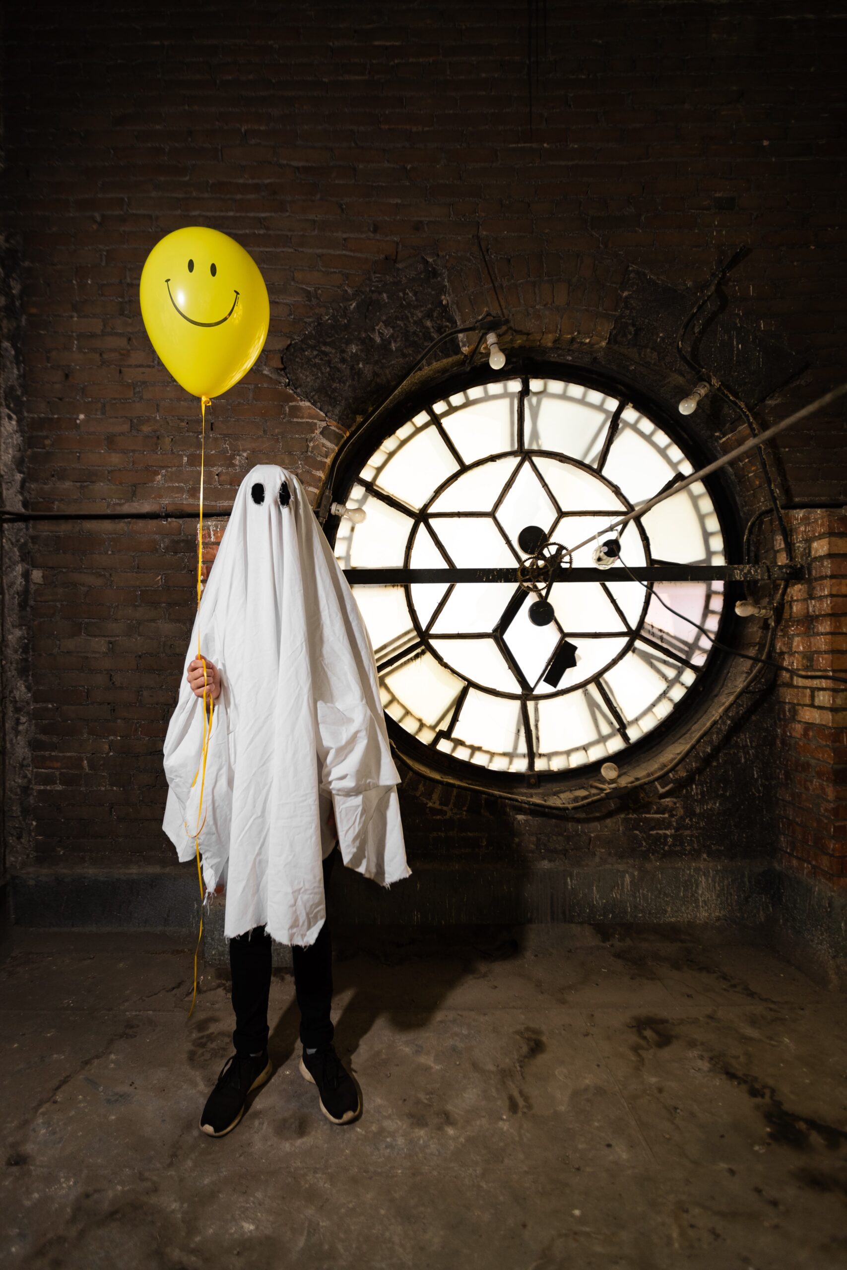 Person in white sheet ghost costume stands in front of large glass clock while holding a yellow balloon with a smiley face