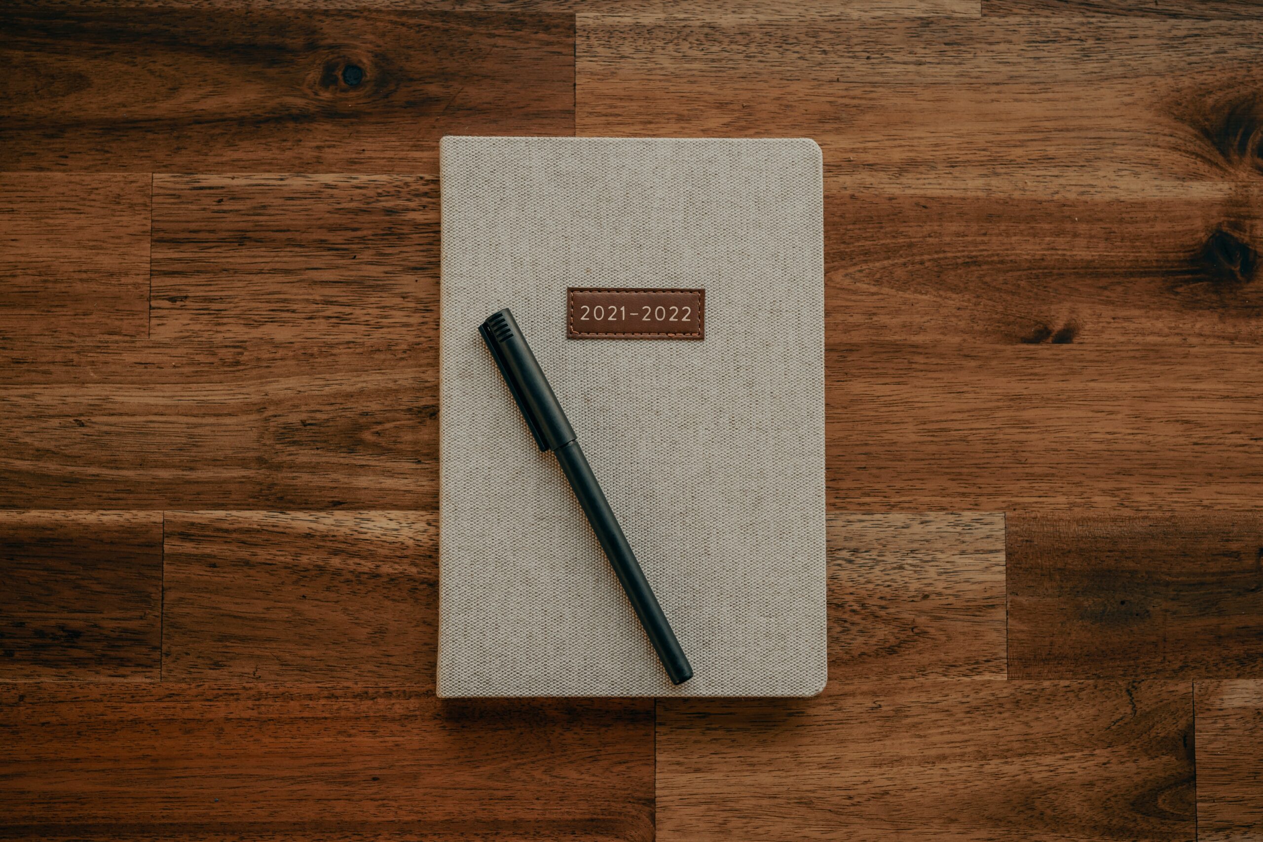 Image is of a beige 2022 day planner with a black pen on top. The planner sits on a dark brown wooden table. This image represents resolutions and habit formation in 2022.