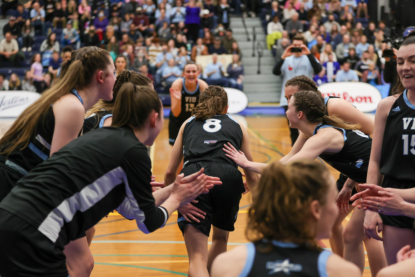 A basketball player in a black uniform runs onto a court to high five a teammate while surrounded by other players applauding her.