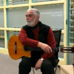 Dr. Tony Robertson, an older man with grey hair and beard, sits with his guitar.